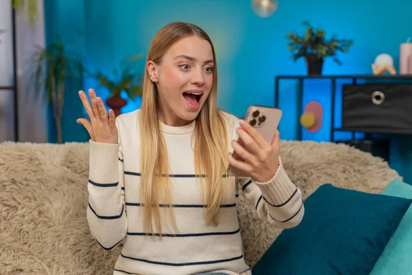 Beyaz bir kadın evdeki kanepede oturmuş online oyunu kutluyor ve zaferi kazanıyor. Heyecanlı kız akıllı telefonu tutuyor, sevinçle el kaldırıyor, gülümsüyor, mutluluğu, heyecanı ve başarıyı içeride hissediyor.