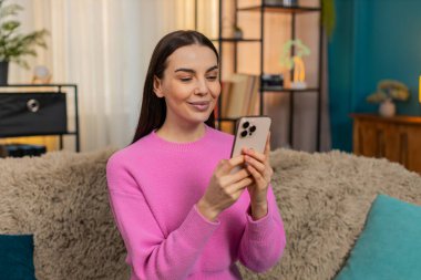 Genç mutlu kadın evinde oturuyor, cep telefonuyla sosyal medyayı karıştırıyor, birden neşe ve şaşkınlıkla gülümsüyor. Kafkasyalı kız kanepede dinleniyor, eğleniyor, neşeli, mutlu, eğleniyor.