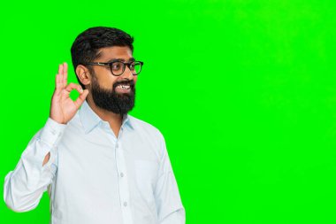 Hintli adam neşeli bir gülümsemeyle iyi el hareketi yapar. Anlaşmayı ve dostça enerjiyle olumlu tutumu ifade eder. Yeşil krom anahtar arka planında izole edilmiş Arap adam onay ve destek gösteriyor