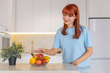 Bardan elma toplayan, meyve kasesinden elma toplayan, ısıran, gülümseyen genç bir kadın. Mutfaktaki kızıl saçlı kız taşınıyor. Mutlu adımlar. Taze meyve atıştırmalığıyla.