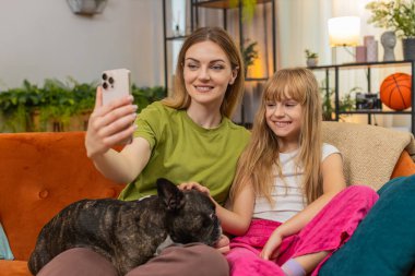 Evde Fransız bulldog 'u olan anne ve kız akıllı telefon şakası gülüşünde selfie filtre maskesi deniyor. Kanepede köpekli kadın ve kız eğlenceli fotoğraf çekiyorlar. Maskelerle oynuyorlar. Sıcak bir anı paylaşıyorlar.