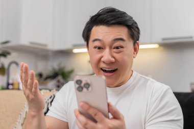 Evdeki Asyalı adam akıllı telefondan iyi haberler okuyor. Vay canına, gözleri kocaman gülüyor ve hızlı cevap yazmaya başlıyor. Kanepedeki Çinli adam sohbette güncellemeleri paylaşıyor. Teşekkürler. Kalp emojisi rahatlıyor ve umut veriyor.