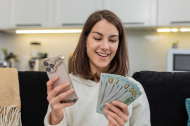 Evde genç bir kadın telefon uygulamalarında nakit para sayıyor gülümseme ve büyük satın alma hedefleri planlıyor. Kanepede oturan beyaz kız maaş ve ikramiye planlarından memnun. Yatırım para tasarrufu ve bütçeden tasarruf sağlıyor.