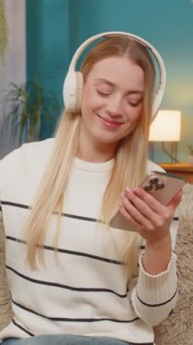 Beyaz, mutlu, genç bir kadın evdeki kanepede oturmuş kulaklıkla müzik dinlemekten zevk alıyor, akıllı telefon tutuyor, gözlerini kapıyor, gülümsüyor, kafasından ritme göre hareket ederek rahatlıyor. İç mekan yaşam tarzı