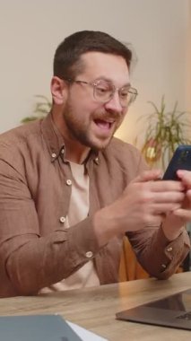 Mutlu genç işadamı akıllı telefon daktilo kullanarak evet diyor, zaferi kutluyor, piyango hedefini kazanıyor olumlu haberler, zafer. Evdeki adam masada oturuyor..