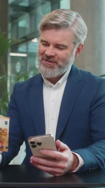 Başarılı, mutlu, olgun bir iş adamı akıllı telefon tutuyor ve ofis lobisinde oturmuş euro banknotlarını sayıyor. İnternette piyango kazanan beyaz sakallı adam. Bütçe hesaplanıyor.