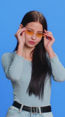 Sarı güneş gözlüğü takan beyaz tenli, şık, havalı bir kadın. Sevimli, neşeli bir gülümseme. Zaferi kutluyoruz. Genç ve mutlu bir kız, mavi arka planda kapalı kalarak heyecandan tatmin olur.