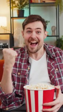 Mutlu Kafkasyalı adam patlamış mısır yiyor, TV seyrediyor, bağırıyor, başarıyı kutluyor, gol zaferini kazanıyor oturma odasında kanepede oturuyor. Heyecanlı genç adam kameraya bakarak yumruklarını sıkıyor.