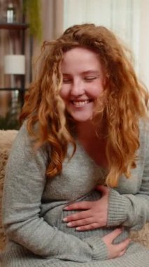 Hamile genç bir kadın kanepesinde oturuyor, karnına hafifçe dokunuyor, gülümsüyor, pozitif havayı kucaklıyor. Kız mutluluğu ve bağlantıyı hisseder, anneliğin küçük anlarında mutluluğu bulur.