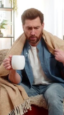 Evdeki genç adam sıcak bir battaniyeye sarılı olarak kanepede oturuyor, kendini daha iyi hissetmek için elinde bir fincan çay tutuyor. Kafkasyalı adam iyi görünmüyor, masada ilaç var, muhtemelen hastalıktan ya da soğuktan iyileşiyor.