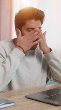 Genç Hintli adam dizüstü bilgisayarda çalışıyor, yorgun görünüyor, evdeki oturma odasındaki kanepede otururken gözlerini ovuşturuyor. İspanyol bir işkolik, görevine devam ediyor, bitkin görünüyor..