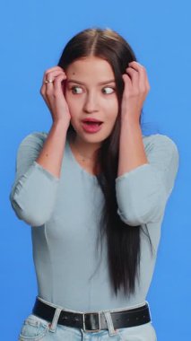 Kafaya dokunan ve patlama gösteren heyecanlı bir kadın endişeli ve şok olmuş görünüyor, başarı fikrini kutluyor. Kazandığına şaşırmış gibisin. Esmer kız, mavi stüdyo arka planında tek başına.