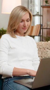 Orta yaşlı bir kadın, dizüstü bilgisayardaki görevi başarıyla bitirdikten sonra sevinçle evinde oturuyor. Yetişkin kadın iş başarısını, gururlu ve olumlu duygusal ifadeyle gülümseyerek kutluyor..