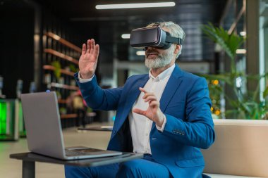 Olgun bir iş adamı yazılım geliştiricisi kulaklık uygulaması kullanarak sunum yapıyor. Sanal gerçeklik 3D 360 videosu izliyoruz. Ofis lobisinde çalışan, VR gözlüklü, beyaz saçlı, takım elbiseli adam..