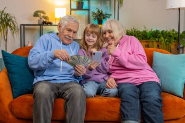 Beyaz büyükbaba, büyükanne ve torun evde akıllı telefon hesap makinesi kullanarak para sayıyorlar. Gülümseyen heyecanlı kız ve yaşlı büyükanne ve büyükbaba koltukta oturmuş piyango kazanmayı kutluyorlar.