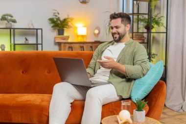 Kahverengi saçlı ve web kamerasıyla konuşan mutlu genç adam uzaktan meslektaşlarıyla çevrimiçi video konferans görüşmesi yapıyor. Beyaz yetişkin bir adam evdeki oturma odasındaki kanepede dizüstü bilgisayar kullanıyor..