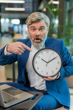 Senin sıran. Diz üstü bilgisayarı olan yetişkin bir iş adamı duvar saatini gösteriyor, onaylıyor, işaret ediyor, işe başlama zamanı, son teslim tarihi. Ofis lobisinde oturan beyaz takım elbiseli adam kameraya bakıyor.