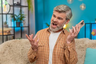 Evde koltukta oturan olgun bir adam hoparlörde akıllı telefon tutarak stres gösteriyor. Orta yaşlı bir adam sinirli görünüyor. Telefon kavgası sırasında el hareketi yapıyor. Olumsuz duygular.