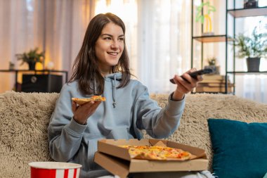 Lezzetli abur cubur pizza yiyen ve patlamış mısır yiyen genç beyaz bir kadın kanepede oturup televizyon ya da oturma odasında film seyrediyor. Günlük kıyafetlerle gülümseyen kız dairede hafta sonunun tadını çıkarıyor.