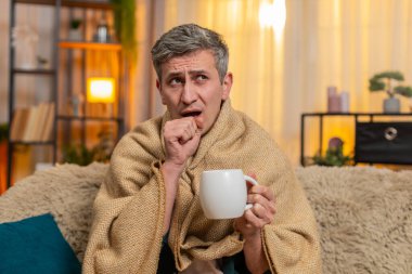 Evdeki üzgün genç adam bir fincan çayla battaniyeye sarılmış, hasta görünüyor ve öksürüyor. Kanepedeki beyaz adam soğuk algınlığı belirtileri gösteriyor, sessizce dinleniyor ve hafif bir hastalık ve yorgunluktan kurtulmaya çalışıyor.