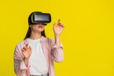 VR kulaklık takan genç bir kadın ellerini havaya kaldırıyor ve sanal dünyaya hayran kalıyor. Sarı arka planda izole edilmiş bir kız dijital deneyime olan hayranlığını ve heyecanını ifade ediyor.