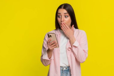 Genç bir kadın akıllı telefon ekranına bakarken şaşırdı, beklenmedik sürprizlere şok etkisi yarattı. Sarı arka planda izole edilmiş bir kız şaşırtıcı içerikten hayret verici bir duygusal ifade sergiliyor..