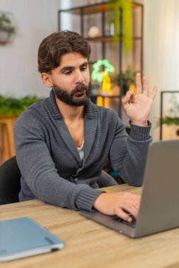 Evdeki Hintli adam dizüstü bilgisayardaki detaylar dijital kontrat dolduruyor elektronik imza ekliyor nefes alıp veriyor. Masadaki Arap adam onaylanmış el işareti gülümseme sakin ruh hali görev onaylandı