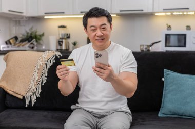 Asyalı adam ödemeyi akıllı telefon ve kredi kartıyla tamamlıyor ve onaylandıktan sonra gülümsüyor. Kanepedeki Çinli adam onaylı tabelayı paylaşırken sohbet güncellemesi yaptı. Faturayı sakladı ve rahatlattı..