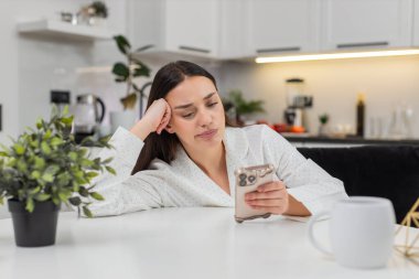 Akıllı telefondaki genç kadın, mutsuz, düşük enerjili, duygusal tükenmişlik halinden sıkılmış. Kanepede oturan beyaz kız beslenme zamanını okuyor. Başparmak hareket ediyor. Yorgun gözler kırılıyor..