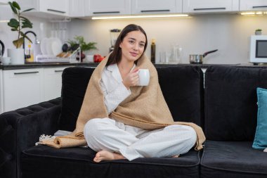 Evdeki genç bayan battaniyeye sarılı çay içiyor rahatla nefes al nazikçe ara ver. Kanepede oturan beyaz kız elinde kadehini yavaşça yudumluyor gözlerini kapatıyor rahat dinleniyor yumuşak gülücükler ılık ruh hali sessiz akşam havası..