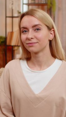 Genç yetişkin bir kadın, evinde kanepede otururken içten ve parlak bir gülümsemeyle kameraya bakar ve kendine güveni yayar. Sarışın kız neşeli duygularını ifade ediyor, mutluluk ve iyimserlik havası yaratıyor.