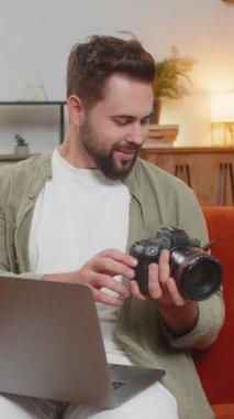 Kanepede, elinde dijital kamerayla, sınıftaki dizüstü bilgisayarın önünde otururken kurgu için fotoğraf seçen mutlu beyaz erkek fotoğrafçı portresi..