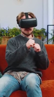 Sakallı adam sanal gerçeklik teknolojisi VR uygulama kulaklığı kullanarak 3D 360 çevrimiçi video oyunu simülasyonu yapıyor ve modern ev dairesinde film izliyor. Gözlüklü adam kanepede oturuyor.