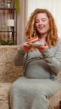 Hamile, genç, mutlu bir kadın elinde donutla kanepede oturuyor. Neşeli bir ifadeyle gülüyor, tatlı bir zevkin tadını çıkarıyor. Kız küçük zevkleri kucaklıyor, mutluluğu basit ve lezzetli anlarda buluyor..