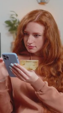 Kredi kartı ve akıllı telefon kullanan mutlu, kızıl saçlı bir kadın para transfer ederken internetten alışveriş nakitsiz yemek siparişi veriyor. Kanepede oturan genç kız.