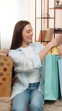 Genç bir kadın alışveriş çantasından aldığı yeni kıyafetleri mutlulukla açıyor. Kanepede oturan beyaz bir kız alışveriş sonrası heyecanlı, şık bir moda ürününden memnun.