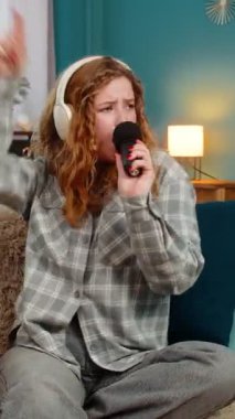 Evde kulaklık takan genç bir kadın mikrofona duygusal bir şekilde şarkı söylüyor ve yüksek sesli müzik ritminin tadını çıkarıyor. Kafkasyalı kızıl saçlı kız sevinçle dans ediyor, kendini sahne yıldızı gibi hissediyor, enerji dolu.