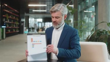 Olgun Kafkasyalı patron iş adamı çevrimiçi video sunumu yapıyor, grafik analizleri gösteriyor, planlama bütçesi, istatistik. Ofis lobisinde yakışıklı bir adam kameraya bakıyor, konuşuyor..
