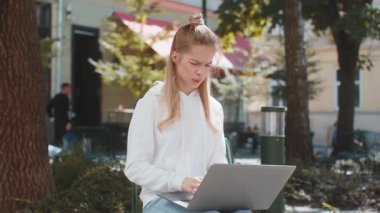 Laptop kullanan, kameraya bakan ve şehir sokaklarında sandalyeye otururken baş parmağını öne eğen genç bir kadın. Kafkas kız olumsuz duygularını ifade eden bir defter üzerinde çalışıyor..