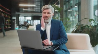 Ofis lobisindeki beyaz iş adamı dizüstü bilgisayarla çalışıyor ve diş ağrısının elinle yanağa değdiğini hissediyor diş hastalığından dolayı acı veren bir ifade. Pulpitis, sakız hastalığı, diş hastalıkları konsepti.