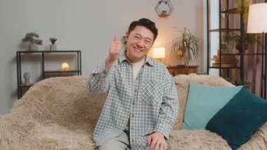 Heyecanlı gülümseyen Çinli adamın portresi onaylayıcı bir şekilde kameraya bakıyor işaret gibi, iyi haber, olumlu geri bildirim gibi. Mutlu Orta Doğulu adam evdeki oturma odasında kanepede oturuyor.