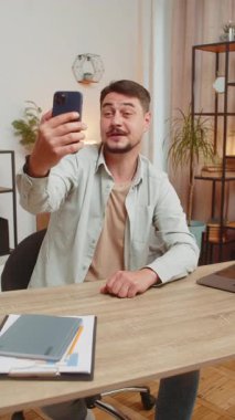 Kafkasyalı serbest çalışan genç adam sosyal medya için akıllı telefon görüntülerini kullanıyor. Masada oturmuş gülümsüyor ve ev ofisinden çalışıyor. Erkek öğrenci çalışması, e-öğrenme, uzaktan kumanda. Dikey.