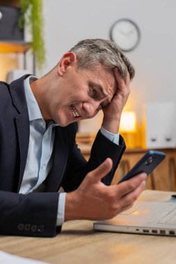 Üzgün genç işadamı, ofis masasında akıllı telefondan kötü haberler okuyor. Resmi takım elbiseli üzgün erkek işveren hoş olmayan tebligattan rahatsız, internetteki bilgiden şok olmuş, mesajla depresyona girmiş..