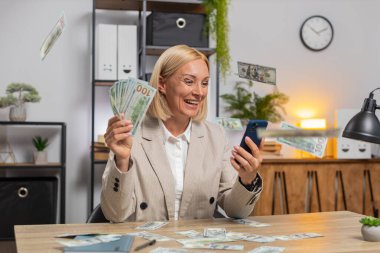 Yetişkin bir iş kadınının üzerine yağmur gibi para yağıyor. Akıllı telefon kullanarak başarıyı kutluyoruz. Oyun piyangosunu kazanıyoruz. Kız ofis masasında oturuyor..