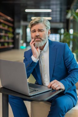 Ofis lobisindeki beyaz iş adamı dizüstü bilgisayarla çalışıyor ve diş ağrısının elinle yanağa değdiğini hissediyor diş hastalığından dolayı acı veren bir ifade. Pulpitis, sakız hastalığı, diş hastalıkları konsepti.