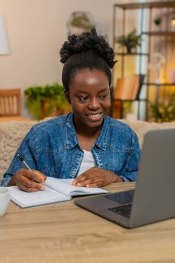 Afro-Amerikalı genç bir kadın evdeki kanepede otururken dizüstü bilgisayarda ders çalışıyor. Siyah kız not defterine notlar alır. Odaklanmış, özenli ve uzaktan öğrenme sürecine odaklanmış görünür.