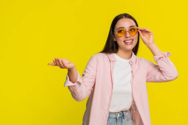 Şık bir güneş gözlüğü deneyen genç bir kadın neşeli bir ruh hali sergileyen komik bir ifadeyle neşe içinde poz veriyor. Sarı arka planda izole edilmiş bir kız neşeli bir enerji gösterisi sergiliyor.