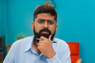 Evindeki Hintli adam kalemini elinde tutuyor. Dikkatlice ileriye bakıyor ve odaklanmış bir zihinle görevler planlıyor. Yakın plan Arap adam bir sonraki hamle ve stratejik fikirler üzerinden kalem çevirme hedefleri düşünüyor