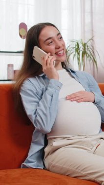 Oturma odasındaki koltukta oturmuş akıllı telefondan konuşan hamile bir kadın. Evinde cep telefonu tutan mutlu bir beyaz kadın. Doğum öncesi bakım ve hamilelik konsepti.