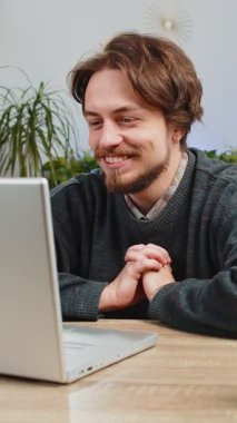 Beyaz bir adam kanepeye oturmuş kameraya bakıyor arkadaşlarıyla ya da ailesiyle görüntülü bir konferans görüşmesi yapıyor. Ofis masasında el sallayarak gülen adam.